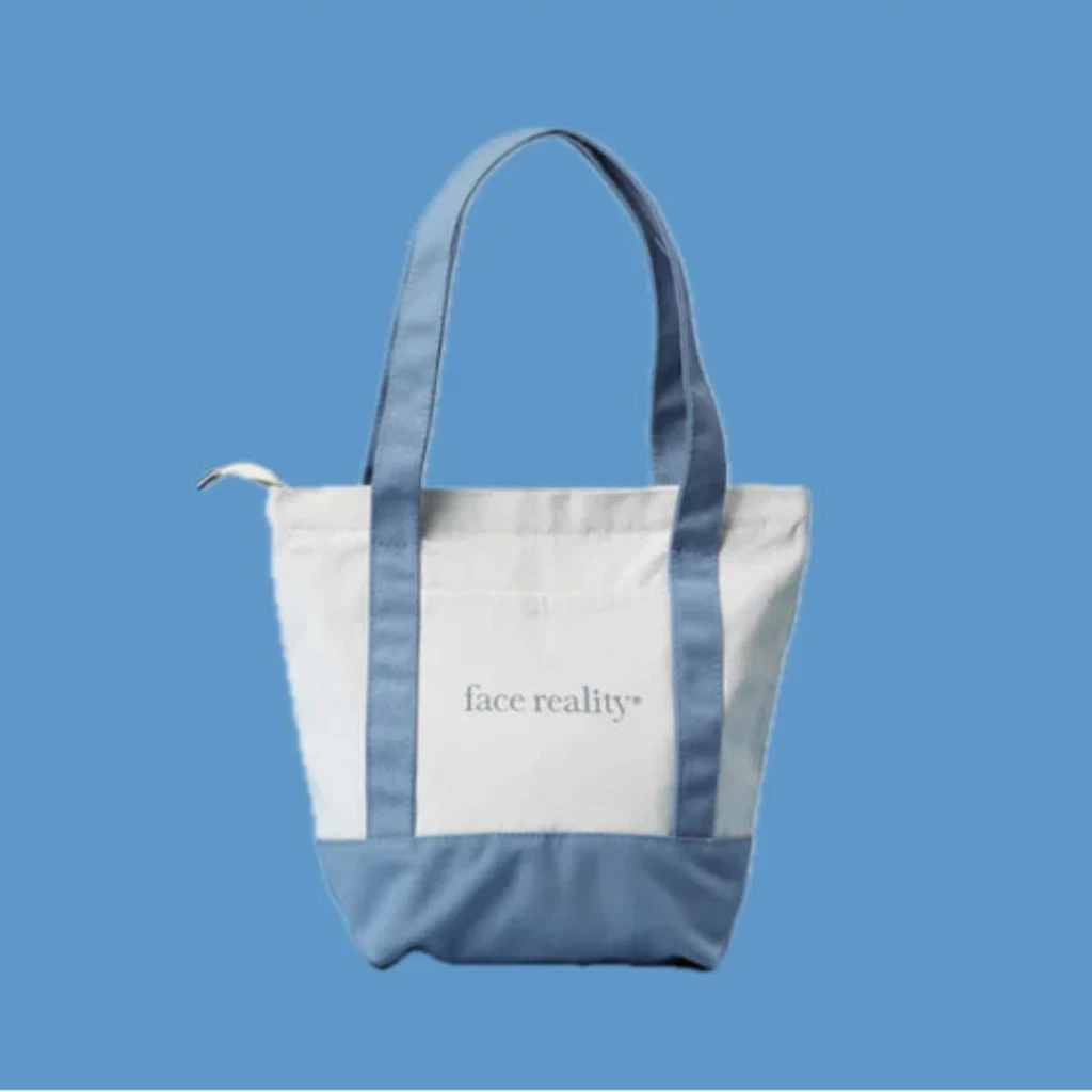 A white canvas tote bag with light blue handles and bottom panels, featuring the text 'face reality' on the front. The bag is centered against a solid blue background