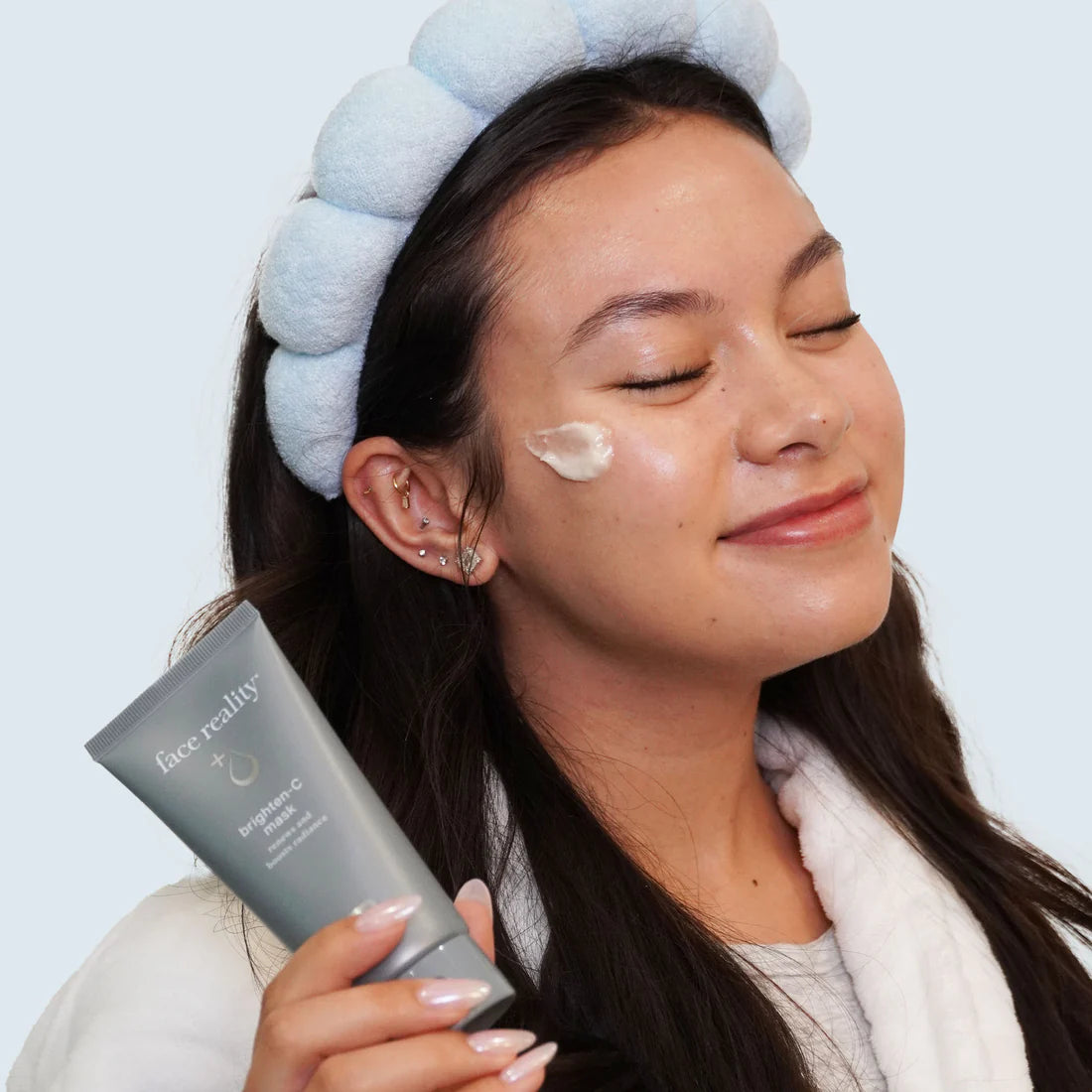 Woman applying cream to her face with a blue towel on her head, holding a skincare product.