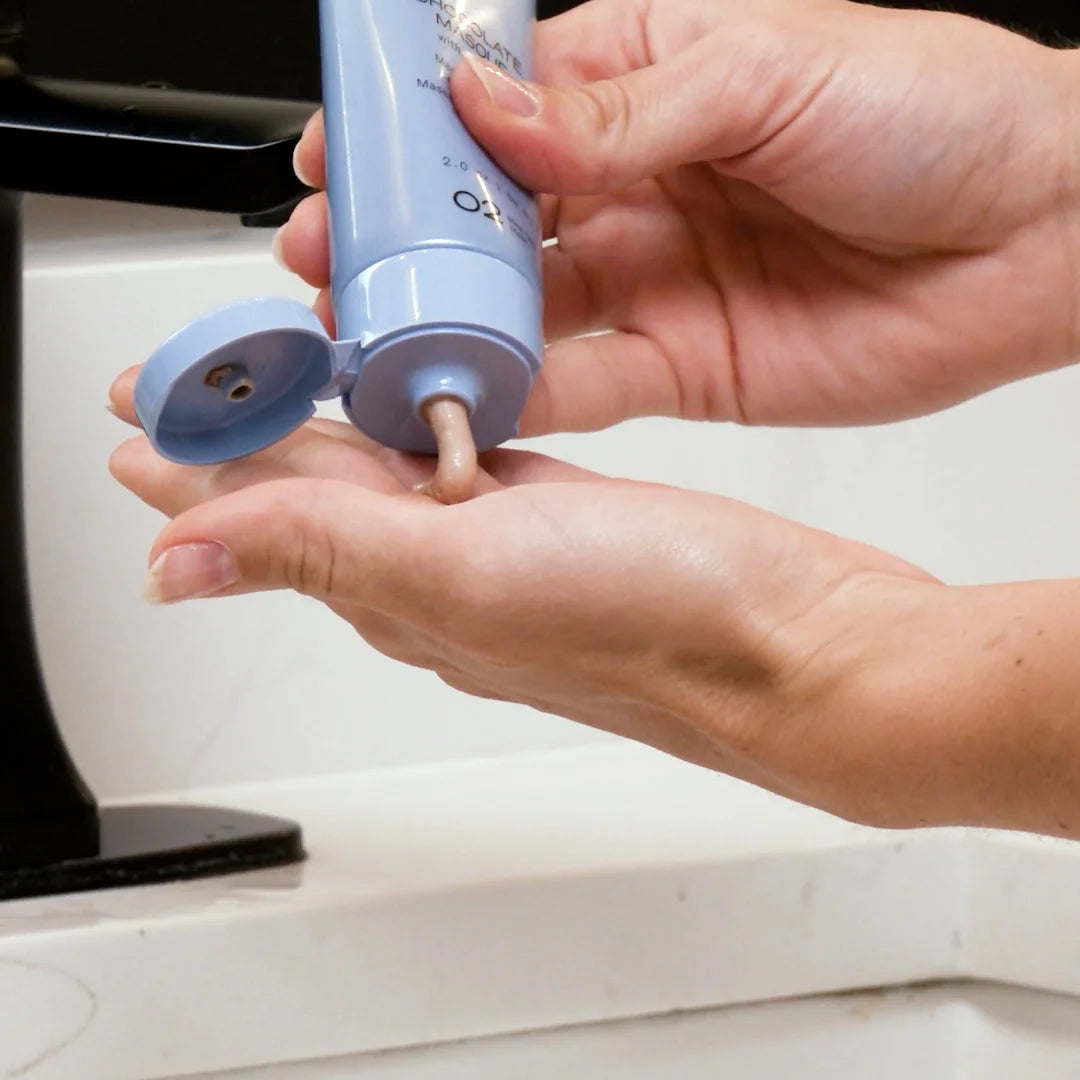 Hand applying a tube of cream to another hand against a neutral background