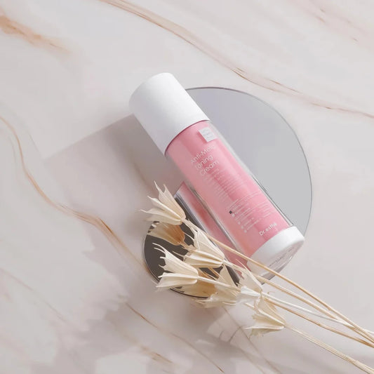 A pink dr esthe anti mela toning cream bottle with a white cap, placed diagonally on a round mirror over a light marble surface with dried flowers in the foreground.