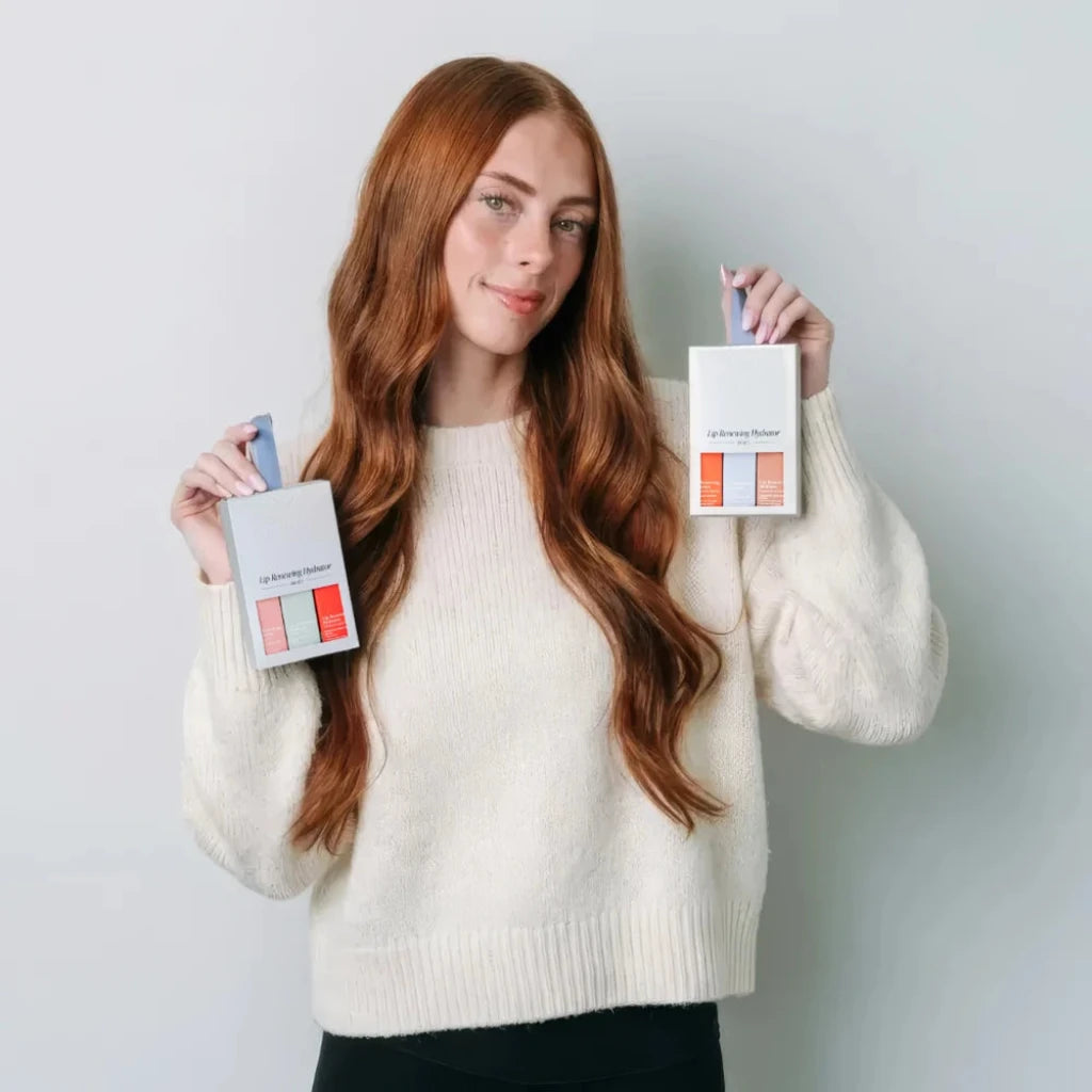 A woman with long red hair holds up two silver boxes of Circadia Lip Hydrator gift sets, one showing coral and pink shades and the other showing peach and orange shades, against a light gray background.