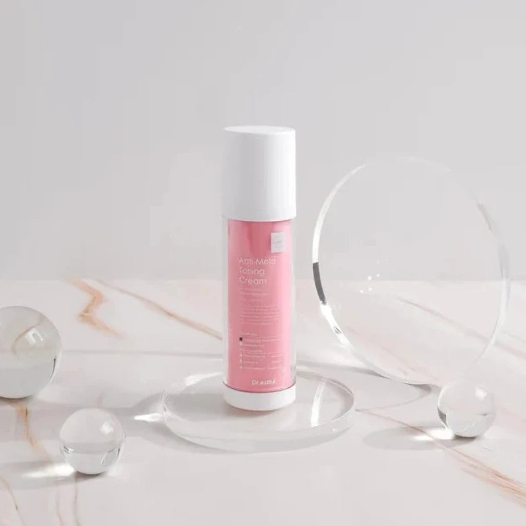 A pink and white Dr Esthe Anti Mela Toning Cream bottle stands upright on a clear acrylic platform on a light marble surface, with a large clear circular prop behind it and small clear spheres around it.