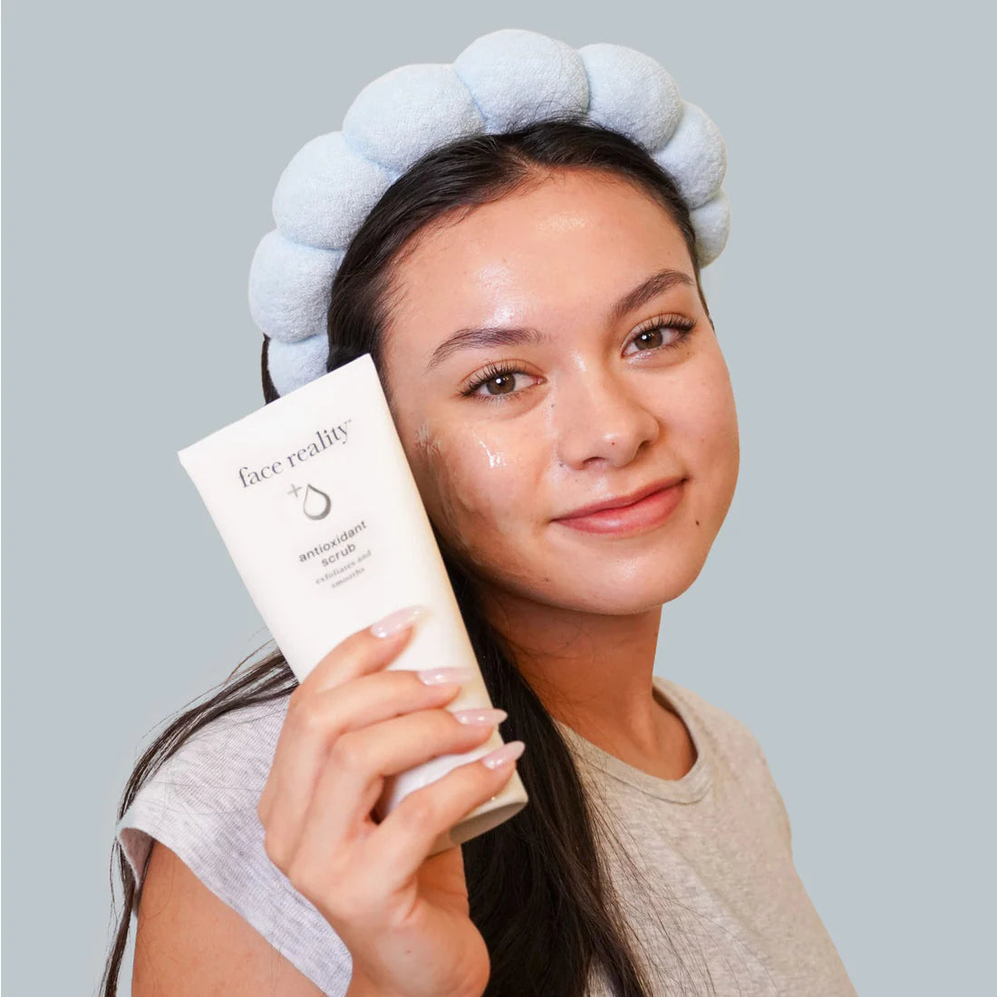 Woman applying facial product with a gray towel on her head against a plain background