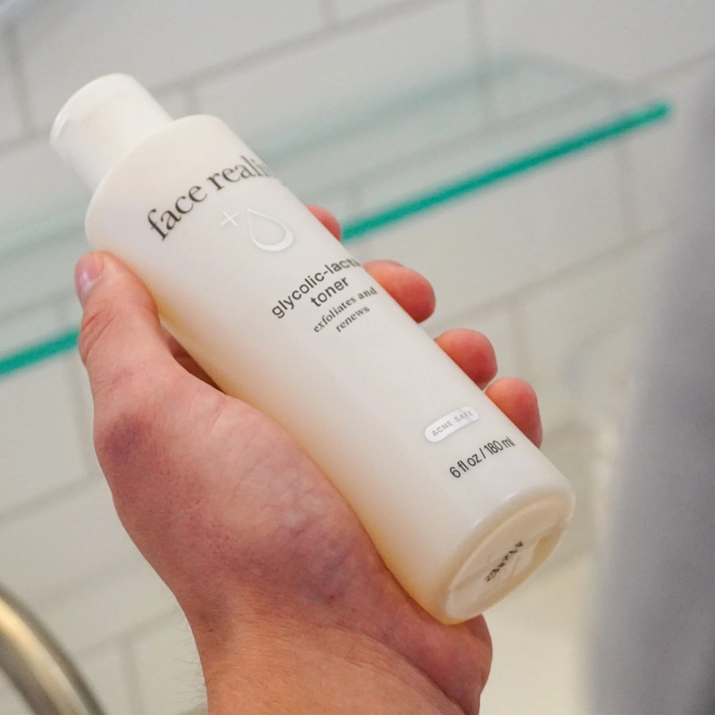 Hand holding a bottle of 'face reality' glycolic acid toner in a bathroom setting.