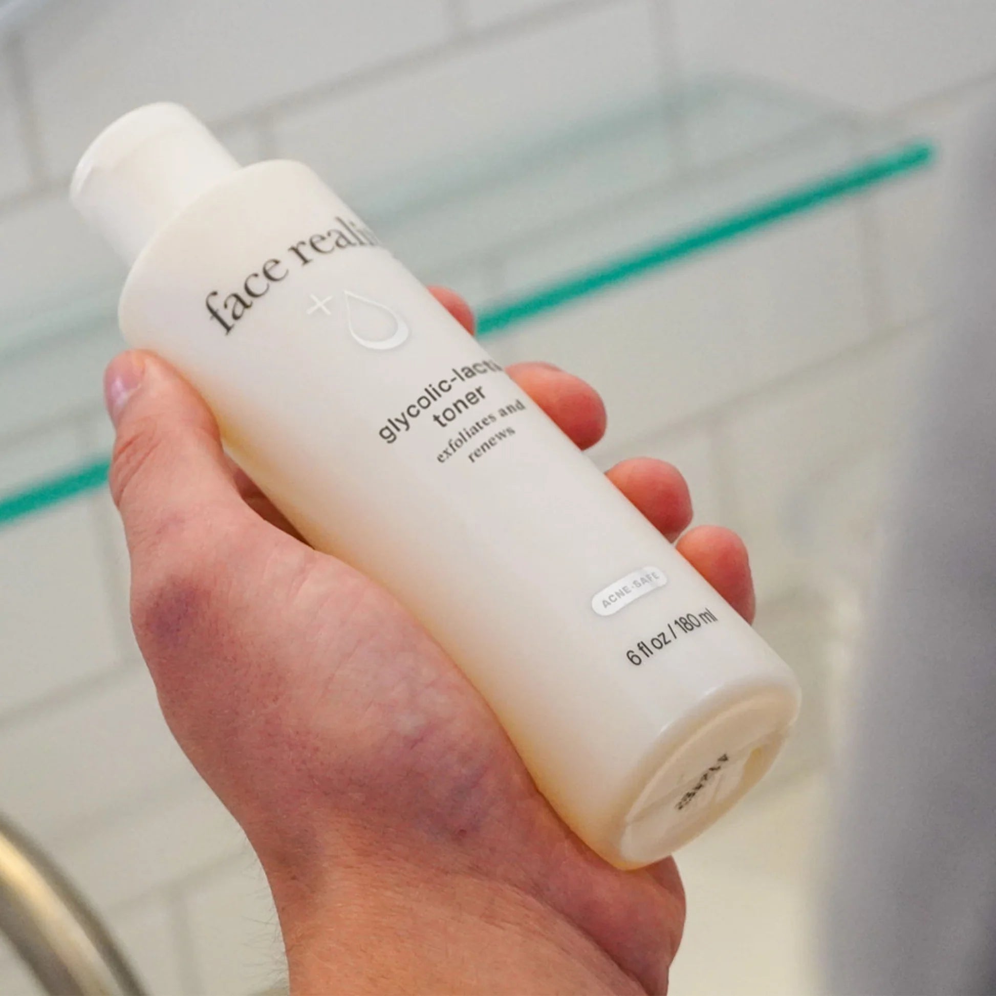 Hand holding a bottle of 'face reality' glycolic acid toner in a bathroom setting.