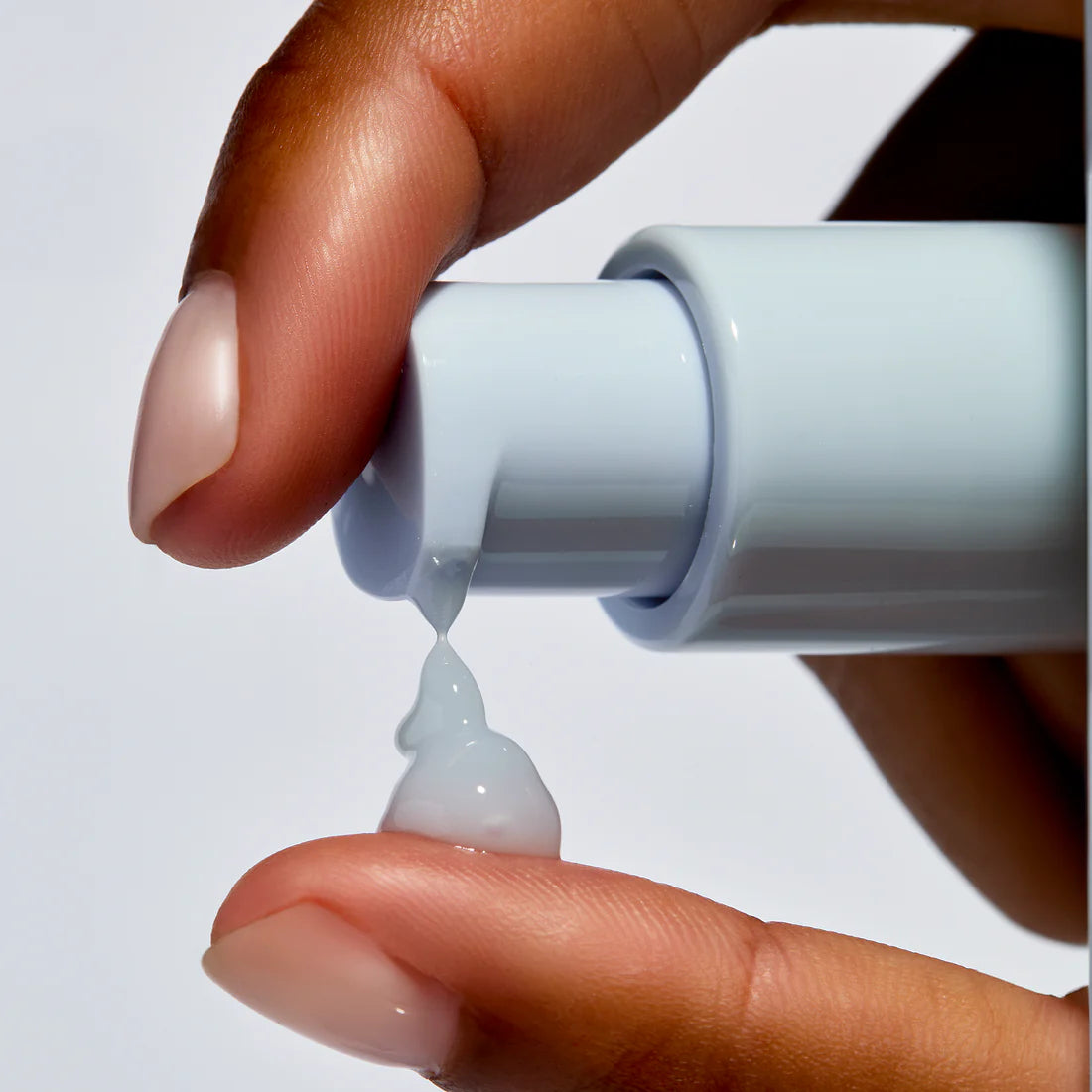 Hand holding a small tube with a white cap, squeezing out a drop of clear gel onto a white background.