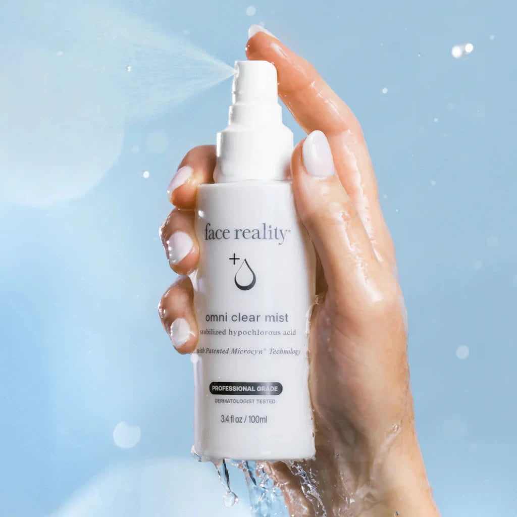 A hand holding a white Face Reality Omni Clear Mist spray bottle with water droplets spraying from the nozzle, set against a light blue background.