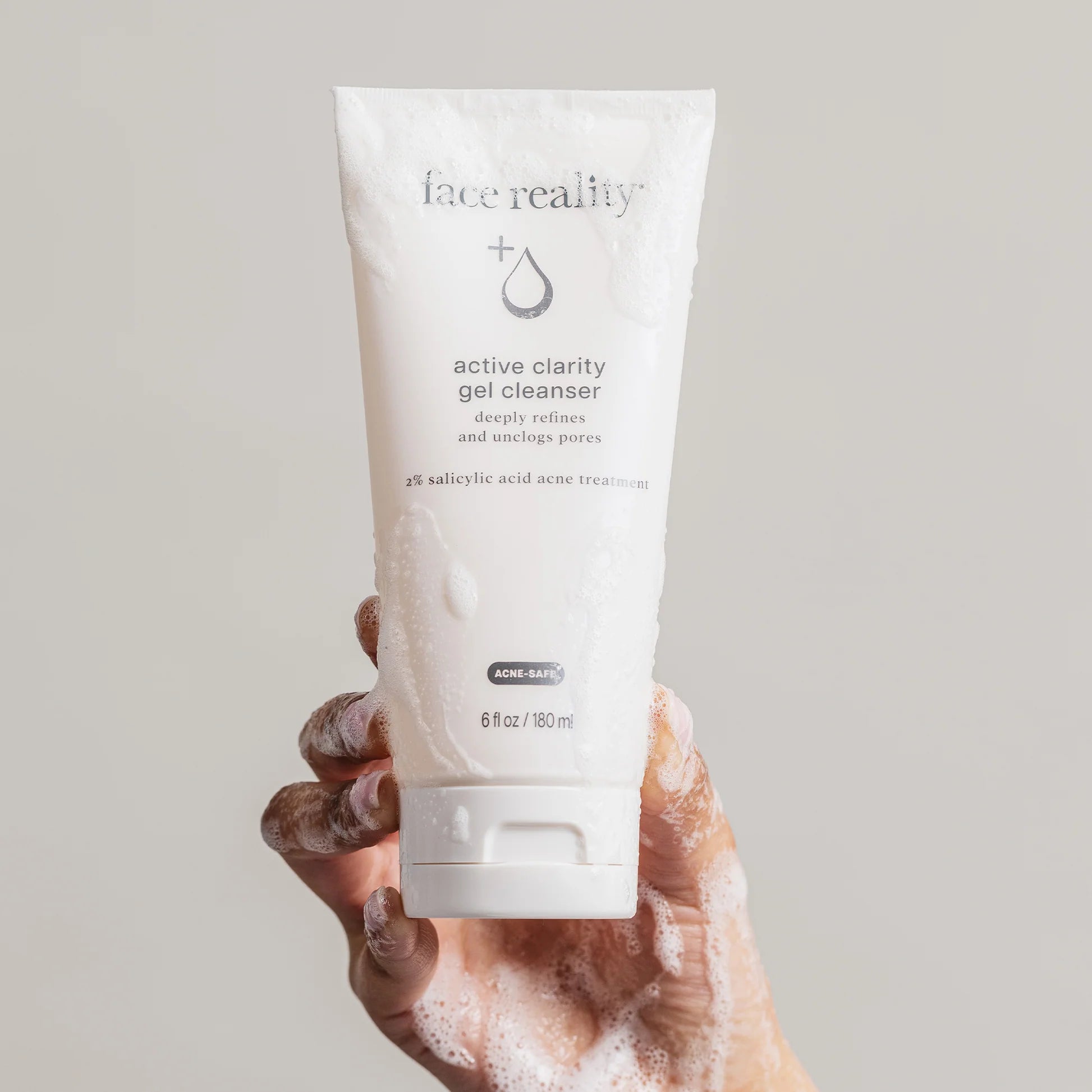 A hand covered in soap lather holds a white squeeze tube of face reality Active Clarity Gel Cleanser against a solid light gray background.