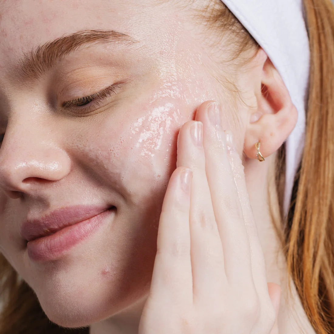 Close-up of a woman with closed eyes applying a glossy, translucent face reality active clarity gel cleanser to her cheek with her finger. She has a white headband on and a small gold hoop earring.
