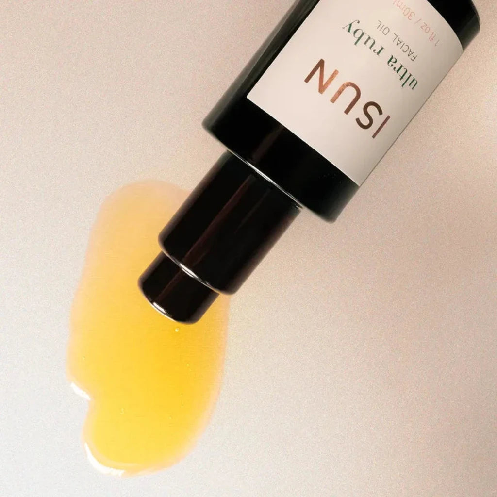 A bottle of ISUN Ultra Ruby Facial Oil lies diagonally on a light beige surface, with a glossy smear of golden yellow oil spread beneath the open cap.