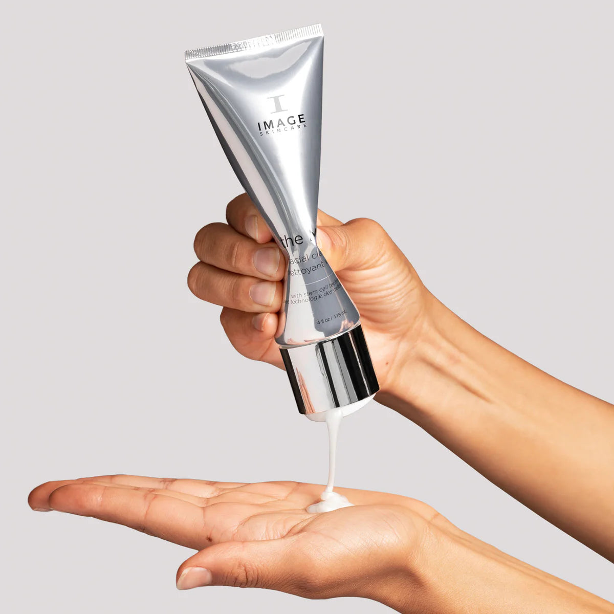 Hand holding a silver skincare tube labeled 'IMAGE' with cream being dispensed onto another hand against a gray background.