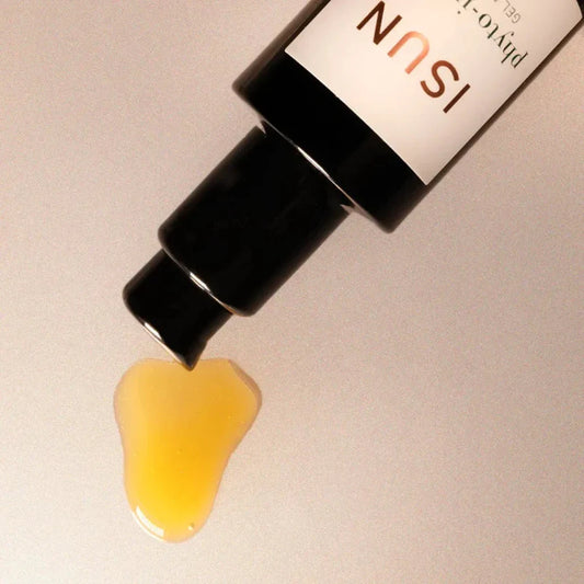 A close-up of a skincare bottle with its black cap unscrewed, dispensing a glossy droplet of yellow gel onto a light beige surface. The bottle has a white label with gold text.
