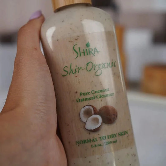 Hand holding a bottle of Shira Shir-Organic Pure Coconut Oatmeal Cleanser with a blurred background.