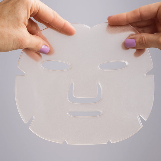 A translucent clear silicone or plastic sheet mask with facial cutouts, held by two hands against a light gray background.