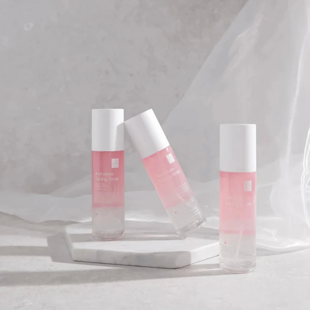 Three pink dr esthe anti mela toning toner  bottles with white caps arranged on a white marble slab against a soft gray textured backdrop.