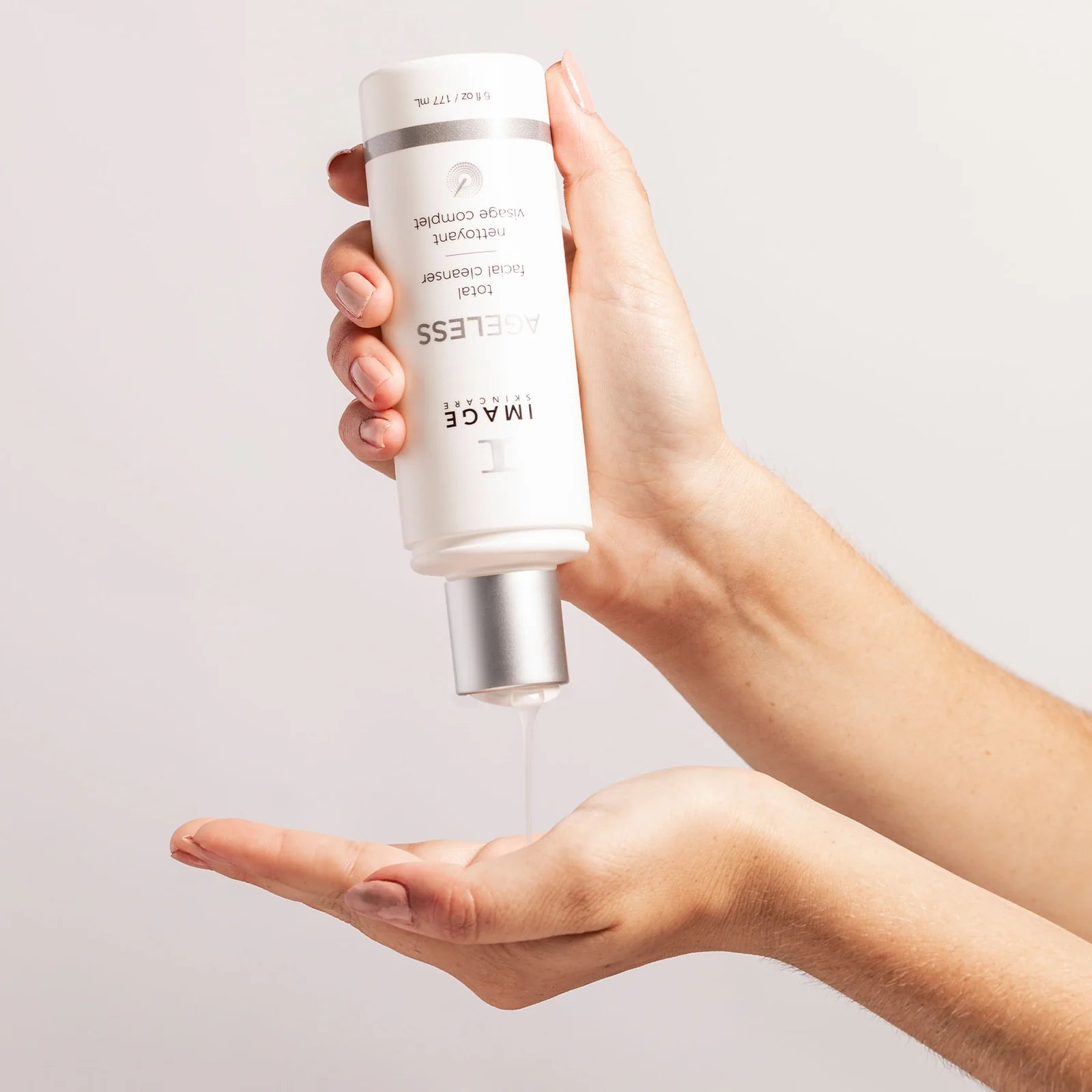 Hand holding a skincare product labeled IMAGE skincare AGELESS total facial cleanser hands pouring liquid against a light background